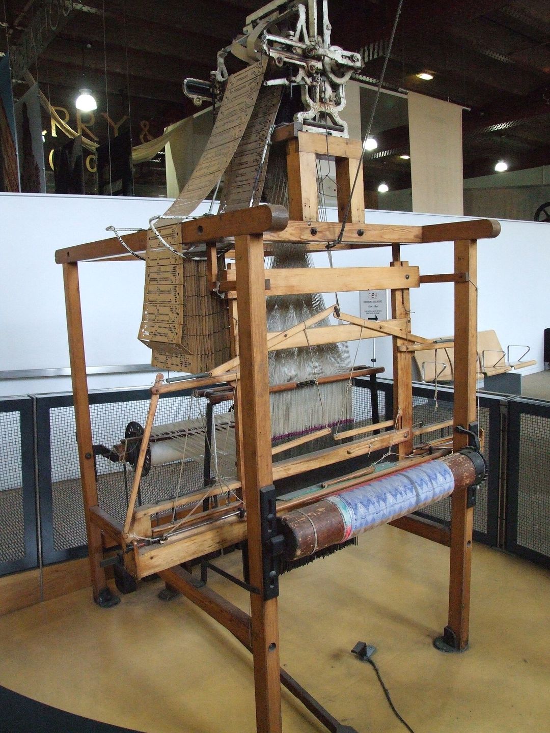 a wooden jacquard loom with its chain of punch cards hanging from the card-reading head above the warp threads